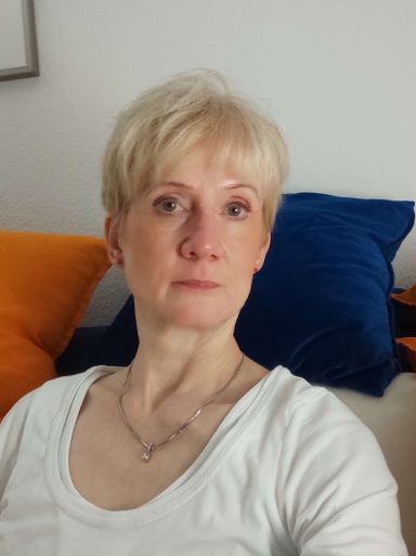 Blonde Frau mittleren Alters, weiß, auf einem Sofa sitzend, schaut in die Kamera